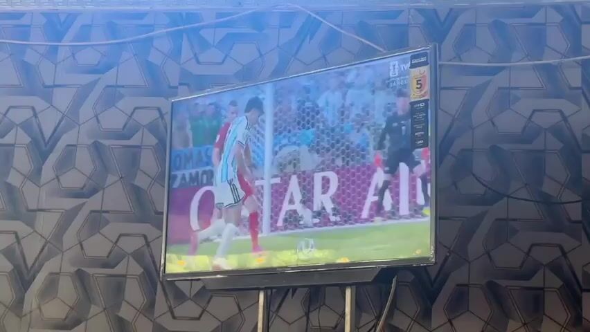 A large television screen displays a soccer match. The screen shows players in red and white uniforms on a green field. In the foreground, a player in a white and blue striped uniform is kicking a soccer ball. Behind the player in red, a goalkeeper in a yellow uniform stands in front of a net. The background of the screen shows a stadium filled with spectators. The television is mounted on a black stand. The wall behind the television has a grey geometric pattern. The lighting in the room is bright, with a blue tint coming from above.