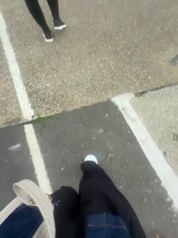 The video opens with a shaky, first-person perspective shot of someone's feet walking on a pavement. The pavement appears to be an outdoor area with white lines marking parking spots. In the background, another person's legs and feet, wearing black leggings and white sneakers, are visible. The camera then pans to the right, revealing a slightly blurry view of a harbor. A boat is docked on the left, with a yellow rope tied to a bollard in the foreground. The water is calm and gray under an overcast sky. Further in the distance, industrial structures with cranes are visible along the waterfront. The scene is a harbor, likely a commercial port, with a boat and industrial equipment.