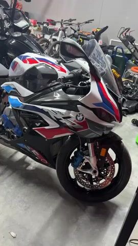 The video shows two motorcycles parked inside a garage. In the foreground is a BMW sport bike with a red, white, and blue color scheme. It has black wheels, blue brake calipers, and a silver exhaust pipe. Behind it is a larger, black motorcycle, possibly a touring bike or cruiser. In the background, there are several bicycles, a lawnmower, and other garage items. The garage has a concrete floor and white walls. The lighting is bright. There are no people visible in the frame.