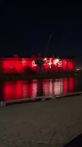 At night, a large, dark brick building is illuminated with bright red light, reflected in the calm water alongside a paved walkway. A crane is affixed to the building, and a projector displays what appears to be a light show on the wall. Across the water, city lights shine in the distance. A person in a white jacket walks along the walkway. Another person walks along the walkway taking pictures.