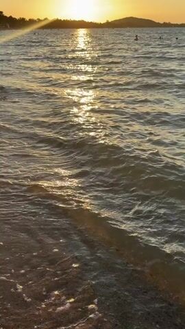 A tranquil ocean scene at sunset. The sun, a bright orb, is low on the horizon, casting a warm golden glow across the water. The water is calm with gentle waves lapping at a sandy shore. In the distance, silhouettes of trees and a small hill can be seen. There are a few people visible in the water, appearing to swim or wade. The overall mood is peaceful and serene.