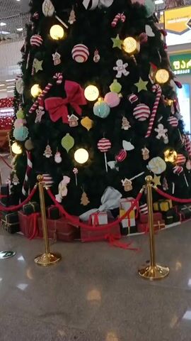 The video shows a large Christmas tree in what appears to be a shopping mall or airport. The tree is decorated with a variety of ornaments, including candy canes, gingerbread men, striped balls, and white bows, and is lit with warm white lights. There are wrapped gifts placed around the base of the tree, which is enclosed by gold stanchions with red rope. The surrounding area is brightly lit, with other shops and decorative elements visible in the background. The camera slowly pans up to show the top of the tree.