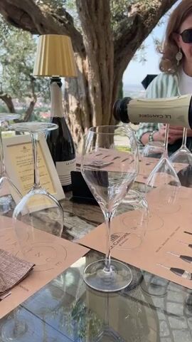 In a mid-shot, a bottle of Le Cantorie sparkling wine is being poured into a tall-stemmed glass on an outdoor table. The table is covered with a glass top and patterned paper placemats, and is set with several wine glasses. A framed card and a small lamp sit on the table, and in the background, a woman in a striped shirt is partially visible. Beyond the table, trees and a distant landscape can be seen under a clear sky.