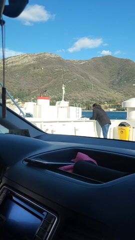 A view from inside a car on a ferry. In the foreground is the dashboard of the car, with a phone visible in a storage compartment. Through the windshield, a person stands on the ferry's deck, leaning against the railing. Behind them, there is a body of water and a mountain range. The sky is blue with scattered clouds. A yellow trash can is visible on the deck next to the person.