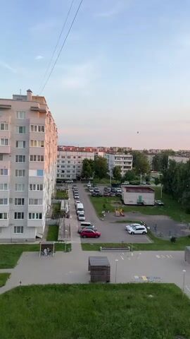 The video shows a panoramic view of a residential area at dusk. The camera pans from left to right, revealing a tall, multi-story apartment building on the left side of the frame. In front of the building, there is a paved courtyard with a playground area and some wooden structures. Several cars are parked along a road that runs through the residential complex. More apartment buildings and trees are visible in the background. The sky is clear with some light clouds, and the setting sun casts a warm glow on the scene, indicating it is evening. There are no people clearly visible in the foreground or engaged in any specific actions. The overall impression is a quiet, ordinary urban neighborhood at the end of the day.