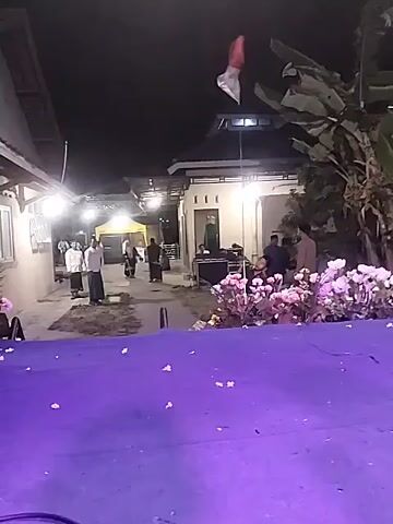 The video shows a nighttime outdoor scene with several people. In the foreground, there is a purple surface, possibly a table or stage covering. Behind this, a walkway or alleyway is visible. On the left side of the walkway, a building with lights is present, and a few people in traditional attire are standing on what appears to be prayer mats, facing away from the camera. In the background, there are more buildings and some trees. To the right of the walkway, there is an area with flowers, sound equipment, and several people. A red and white flag is seen flying and waving in the air. The overall lighting suggests it is dark outside, with artificial lights illuminating the area. The people appear to be engaged in some sort of gathering or event.