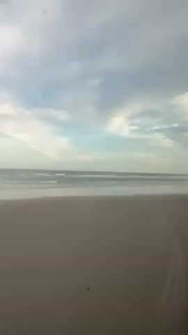 A landscape video shows a mostly flat beach with calm waves coming in. The sky is cloudy with hints of blue peeking through. The camera seems to be at eye level, looking out towards the ocean. There are tire tracks on the sandy beach. No people or objects are visible. The sound of music is playing.