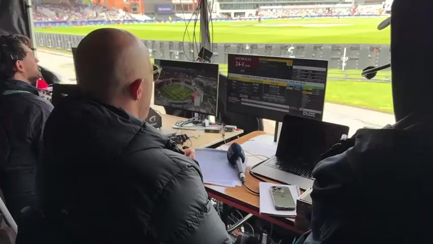 The video shows a behind-the-scenes view of a cricket match from a commentary booth. In the foreground, a bald man wearing glasses and a dark jacket is seated, looking at a monitor. He has a microphone in front of him. To his left, another person wearing a dark jacket is partially visible, also looking towards the monitors. To his right, a person wearing a hooded dark jacket is seen from the back. The booth is equipped with multiple monitors displaying game statistics, a live feed of the stadium, and possibly a laptop. The background reveals a bright green cricket field with players in uniform, a crowd of spectators in the stands, and modern buildings surrounding the stadium. The overall environment is that of a professional sports broadcasting setup during a live event.