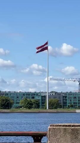 A Latvian flag waves on a tall flagpole against a blue sky with scattered clouds. In the background, there are green trees and modern buildings. A large construction crane is also visible in the distance. In the foreground, a body of water can be seen, with a railing in the lower part of the frame. The camera is positioned at a low angle, looking up at the flag. The video is a short, static shot.