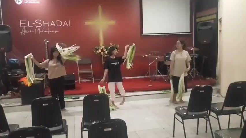 Three women are dancing on a stage in front of a red backdrop with a golden cross and the words "EL-SHADAI Allah Maha Kuasa". The woman on the left is wearing a beige top and black pants, holding green leafy props in each hand. The woman in the middle is wearing a black t-shirt and white pants, holding a bouquet of flowers and green leafy props. The woman on the right is wearing a light-colored top and dark pants, holding green leafy props in each hand. All three women are moving their arms and props in a synchronized dance. There are musical instruments on the stage behind them, including a drum set and a guitar. In the foreground, there are rows of empty black chairs. The lighting is warm, and the overall atmosphere appears to be a performance or worship service.