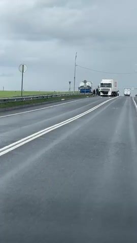 The video shows a wet, gray asphalt road with double white lines dividing the lanes. The camera is positioned low, as though inside a moving vehicle. In the distance, two large trucks are stopped on the side of the road. One is white and the other is blue. There are three people standing between the trucks, seemingly in conversation. Behind the guard rail on the left side of the road is a field of green grass. The sky is overcast with heavy, gray clouds. The road reflects the light from the sky due to the rain. There is a light pole with a traffic sign next to the left side of the road.