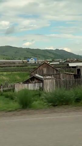 This is a vertical video shot from a moving vehicle, capturing a rural landscape. The foreground shows a blurred asphalt road. Behind the road, there is a strip of green vegetation, also blurred due to motion. The mid-ground features a village with wooden houses, fences, and some trees. The houses appear simple, with some having corrugated metal roofs. Beyond the village, rolling green hills with some trees dominate the background. The sky is cloudy with patches of blue visible. The video captures a sense of passing through a quiet, somewhat undeveloped rural area.