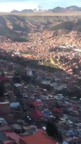 The video shows an aerial view of La Paz, Bolivia. The city is nestled in a valley surrounded by mountains, some of which are snow-capped. The buildings are primarily brick and terra cotta colored, creating a dense urban landscape. A yellow cable car ascends, offering a panoramic perspective of the cityscape.