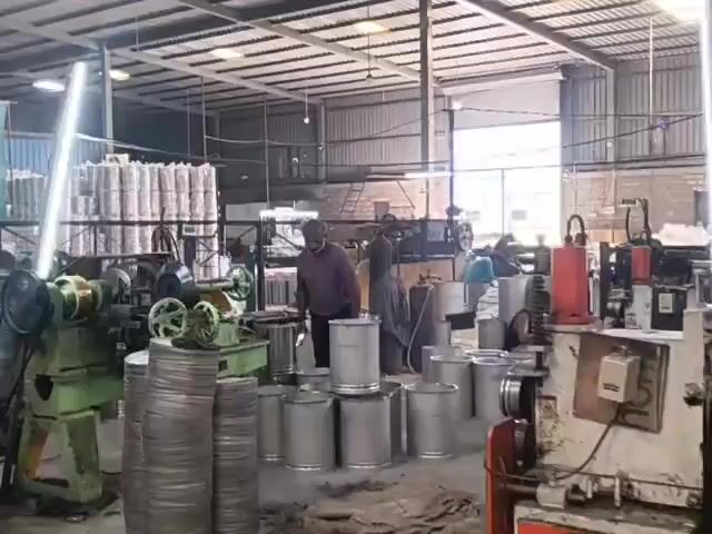 The video displays a busy factory floor. In the foreground, there are stacks of metal cans and machinery. A man is visible in the center, and another man is working on equipment in the background. The factory appears to be involved in the manufacturing or processing of metal containers. The environment is industrial, with overhead lighting and large open spaces. The overall impression is one of active production.