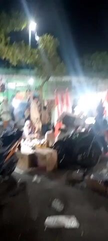The video shows a night scene at an outdoor market. There are several motorcycles, some covered in plastic. People are visible, sitting and standing around stalls displaying various items, including what appear to be household goods and possibly food. Indonesian flags are prominently displayed on poles. The lighting is a mix of streetlights and bright, focused lights, creating a somewhat hazy and dramatic effect. The ground is paved.