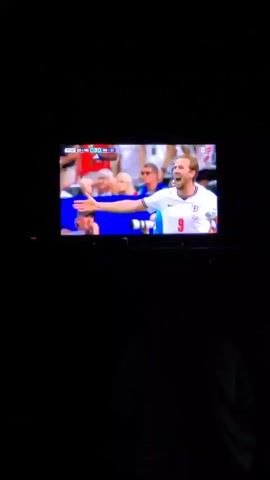 This media file displays a close-up shot of a television screen in a dimly lit room, suggesting it's nighttime. The television is showing a soccer match, with a focus on two players in white jerseys. The score graphic in the upper left corner indicates the time is 73:52 and the score is 0-0. The first player shown is a man with blonde hair, wearing an England jersey with the number 9. He appears to be speaking or shouting, with an animated expression. The second player, seen briefly, is a young man with dark hair, looking intently off-camera. The background of the televised game shows a blurred crowd and stadium elements, indicating a professional sporting event. The setting is a darkened room, with the silhouette of a person visible in the foreground, implying the viewer is watching the match. No weather information is available.