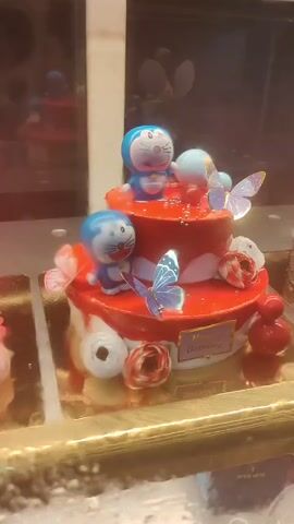 A two-tiered, red-frosted cake is presented in what appears to be a display case. The cake is adorned with two Doraemon figurines, blue and white butterflies, pink and white flowers, and small red decorative elements. A "Happy Birthday" plaque is visible on the lower tier. The background is blurred and shows reflections of other objects, possibly additional cakes or store displays.