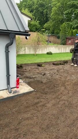 The video captures an outdoor landscaping project in Nový Jáchymov, Czechia, during the daytime, likely under overcast skies or after rain, as the ground appears damp and muddy. The scene is a backyard or garden area where new sod is being laid.

Two men are working. One man, wearing dark clothing and a cap, walks from the right side of the frame carrying a rolled-up piece of sod. He then bends down to place it on the prepared soil. Another man is visible further in the background, near a red and black wheelbarrow, also involved in laying the turf.

In the foreground, a large expanse of tilled, muddy soil is ready for sod. To the left, the corner of a white building with a gray roof and downspout is visible, with a red bottle on a concrete slab nearby. In the mid-ground, sections of green sod have already been unrolled and placed, forming a new lawn. Several other sod rolls are scattered on the ground, awaiting installation. A rake lies on the ground near the newly laid turf.

The background features lush green trees, a light-colored concrete retaining wall, and a wooden fence. A black shipping container is also visible in the background, with a ladder leaning against it and partially visible text such as "20 TDM" and "203134 4" on its side. The overall activity depicts the manual process of transforming a bare soil area into a green lawn.