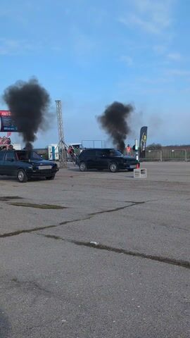 The video shows two vehicles performing burnouts on a paved runway, producing large clouds of black smoke. The cars, a gray Volkswagen Golf and a black hatchback, are lined up side-by-side. After the burnout, they speed down the track, continuing to emit smoke. Spectators can be seen standing behind a fence in the background, observing the event. The sky is blue with scattered clouds. A timing system with lights is visible between the cars at the starting line. The overall setting suggests a drag racing event.