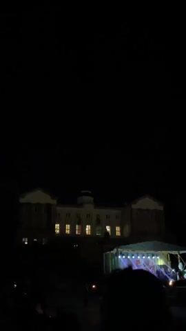 Here's a forensic description of the video:

The video shows a large, multi-story building illuminated at night, with a stage set up in front of it. There is a crowd of people in the foreground. The building appears to have a symmetrical design with a central tower-like structure. Windows are lit up, some showing colored lights. A stage is set up in front of the building with a tent-like covering, with people visible on the stage. There are trees surrounding the building. In the beginning of the video a large firework goes off. The sound is loud.
