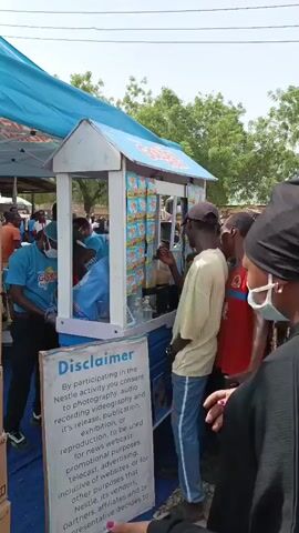 The video shows a crowd of people lining up at a small, covered booth. The booth is painted white and blue, and it has a sign on the side. The sign is titled "Disclaimer" and appears to be related to Nestle. The booth has packages of what looks like Golden Morn breakfast cereal hanging on the front. A person inside the booth, wearing blue, is serving the customers. The people in the line are of various ages and wear different colored clothing. The background shows trees and a bright sky. The atmosphere appears busy and possibly festive. Music plays in the background.