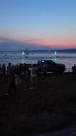 The video captures a sunset scene at a waterfront event. A crowd of people, including adults and children, are gathered on a sandy shore, facing a body of water. Some are holding phones, seemingly recording the event. In the water, several jet skiers or similar watercraft are visible, some with illuminated attachments emitting green lights. One individual is engaged in a water sport, propelled into the air by jets of water, creating a dynamic display against the twilight sky. Sailboats and other vessels are also present on the water, further enhancing the atmosphere of a lively public gathering. The lighting suggests it is dusk, with a colorful sky transitioning from blue to pink and orange hues. A dark-colored pickup truck is parked on the shore behind the crowd, with a metal barrier in front of it.
