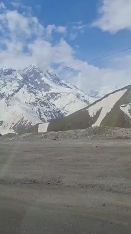 The video shows a landscape of snow-capped mountains on a bright day. The sky is blue with some white clouds. The camera is likely inside a vehicle, as the ground in the foreground looks like a road. The vehicle moves past a metal structure with a partial roof, then a long stone wall. The snow-capped mountains remain visible in the background.