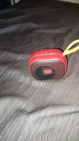 A small, red Bluetooth speaker with a black mesh front and a yellow lanyard sits on a dark gray textured surface. The speaker is positioned at an angle, revealing the mesh and a red center piece. The textured surface appears to be fabric, possibly a bedspread or blanket. The lighting is dim, with the top portion of the scene darker.