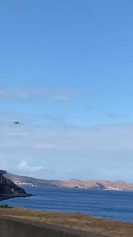A distant airplane is visible in a bright blue sky with scattered clouds. Below the sky is a body of water, likely the ocean, with a distant coastline featuring hills and some buildings. In the foreground, a dark concrete structure is visible, partially obscuring the view. The overall impression is of a scenic coastal landscape on a clear day.