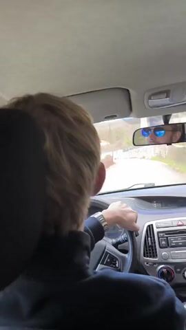 A man with blond hair is driving a car. He is wearing a black shirt and a watch. The camera is behind him, focusing on the back of his head. The car is moving along a road with trees visible outside. Loud music is playing inside the car. The rearview mirror shows the reflection of another person wearing sunglasses.