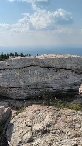 A video captures a large rock face with writing spray-painted in yellow. The writing says "Женя, с юбилеем!" which translates to "Zhenya, happy anniversary!". The rock face is situated on a mountainside with scattered trees and a cloudy sky in the background.