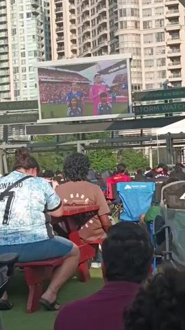 The video shows an outdoor screening of a soccer match on a large screen. A crowd of people are seated in chairs and at picnic tables, facing the screen. The screen displays two soccer players, one in a blue uniform and the other in a pink goalkeeper's jersey, standing with their hands over their hearts, presumably during a national anthem. The people in the foreground are mostly viewed from behind, with one person in a light blue jersey with "RONALDO" visible on the back. The background features tall buildings. The overall atmosphere appears to be relaxed and engaged, as people watch the game together.