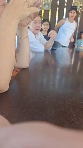 The video shows a group of people sitting and standing around a wooden table. A woman in a white shirt is in the foreground, speaking and gesturing. Behind her, a young girl in a black shirt is looking forward. To the right, a woman in a white tank top is standing and gesturing. Further to the right, a young boy in a grey shirt and a black hat is sitting and looking down. The background consists of a wooden fence with framed pictures. The scene appears to be outdoors, possibly in a cafe or restaurant.