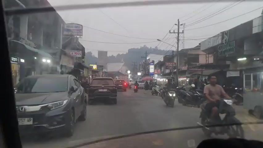 The video shows a street scene with moderate traffic. On the left side, there are several cars parked and moving, including a black SUV and a silver car. The street is lined with buildings, many of which appear to be shops or businesses, indicated by signs such as "RISEKA MAS" and "DANA TUNAI". There are also many motorcycles parked on the right side of the street and a few are moving. People are visible walking and standing on the sidewalks. The sky is overcast, suggesting it might be early morning, late afternoon, or a generally cloudy day. The overall impression is of a bustling urban or semi-urban environment with ongoing commercial activity and traffic.