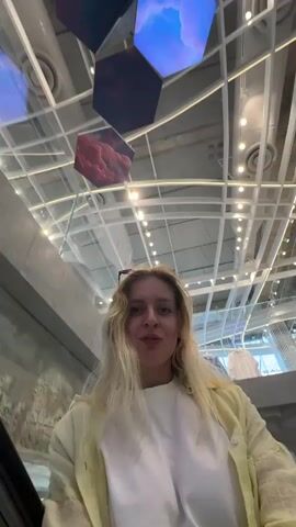 The video shows a woman riding an escalator, looking straight into the camera. The camera is looking up at her from a lower level. She is wearing a white shirt and a light-colored button-down jacket. She has blonde hair. Above her are hexagonal ceiling panels displaying cloud imagery. The escalator is moving upwards, and the background shows an open space with clothing racks and bright lighting. She puts her sunglasses on the top of her head.