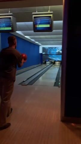 Here is a description of the video:

The video shows a man bowling in an indoor bowling alley. The man is wearing a dark t-shirt and tan pants. He releases an orange bowling ball down the lane toward the pins. The bowling alley has blue walls and a wood floor. There is a screen above the lane displaying the scores. The man bowls and gets a strike.