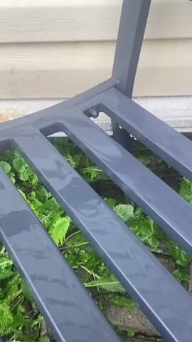 The video shows a close-up view of a wet, gray metal bench. Water droplets are visible on the surface of the bench, indicating it has recently rained or been washed. Green foliage is visible underneath the bench. The background consists of a light-colored wall with horizontal siding. The camera angle is fixed, and there is no discernible movement or action other than the water on the bench.