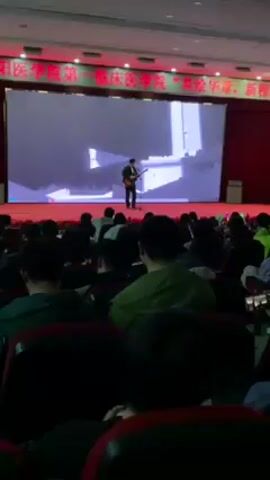 The video captures a scene in a large auditorium filled with seated audience members facing a stage. On the stage, a single individual stands with a guitar, performing in front of a large screen displaying an animated visual. The auditorium is well-lit, with red-cushioned seats and overhead lighting. A red curtain hangs behind the stage screen. The crowd, composed of numerous individuals mostly with dark hair, are seated and oriented toward the stage, indicating they are observing the performance. The performer is positioned centrally on the stage. The atmosphere appears to be one of a presentation or performance, given the stage setup and audience engagement.