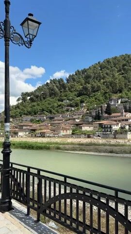 This video captures a scenic panoramic view of the historic city of Berat, Albania, on a clear, sunny day. The camera slowly pans from left to right, showcasing the distinctive architecture of the city. Numerous white, multi-story houses with red-tiled roofs are densely clustered on a steep, verdant hillside, which is covered with lush green trees. In the foreground, a wide, light green river, likely the Osum River, flows calmly. A black metal railing with decorative arches and a black lamppost are visible in the immediate foreground, suggesting the viewpoint is from a bridge or riverside promenade. As the camera pans, a bridge crossing the river and a telecommunications tower on the hilltop come into view. The sky is a bright blue with scattered white clouds. There is no discernible activity other than the camera's movement.