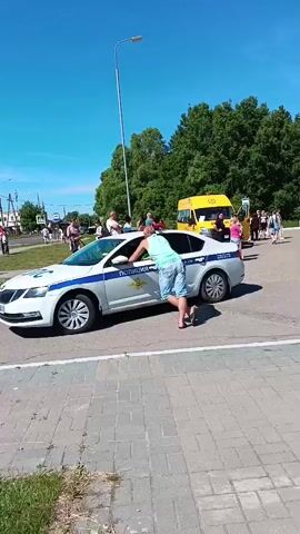 The video opens with a wide shot of a man in a light blue t-shirt and light-colored shorts leaning on the driver's side door of a white and blue police car. The car has "ПОЛИЦИЯ" written on its side. To the right of the police car, there are three yellow buses parked, with people, including children and adults, standing near and boarding them. The bus closest to the police car has "ДЕТИ" written on its side. The background features green trees under a clear blue sky.

The camera then pans to the right, showing more of the paved area where the buses are parked. People are seen milling around the buses, some carrying bags. The camera continues to pan, revealing a white truck with its back open, parked near some benches where more people are gathered. Further to the right, a woman stands next to a baby stroller. In the background, there is a building with blue windows and a white facade. The ground is paved with light-colored bricks.