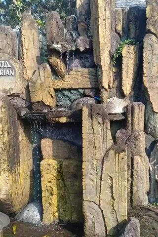 The video shows a close-up, rotating view of a man-made waterfall structure designed to resemble a rock formation. Water flows down over several levels of textured, layered, artificial rocks. The rocks are various shades of brown and yellow, with some appearing darker and more wet where the water has passed. Small green plants are visible growing in some crevices of the rock. In the lower left portion of the frame, a partial sign is visible with the text "RIAN" at the bottom. The background is out of focus, but appears to be foliage and possibly a body of water. The lighting suggests it is daytime outdoors. There are no people or animals clearly visible in the video. The water appears to be clear.