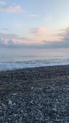 The video shows a pebble beach with waves gently lapping at the shore. The sky is cloudy with a hint of pink and orange near the horizon, suggesting either sunrise or sunset. There are no people visible. The focus is primarily on the natural landscape.