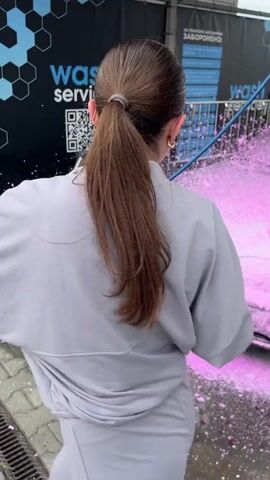 A woman in light grey attire uses a high-pressure washer with a blue and silver nozzle to spray vibrant pink foam onto the front and driver's side of a silver BMW car, which is already substantially covered in the pink substance. The foam splatters on the concrete ground. The scene is set in a car wash bay with a black wall in the background featuring blue hexagonal patterns, white text "wash service," and a QR code.