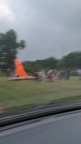A group of people are gathered in a grassy field around a large bonfire. The sky is overcast and appears to be a dull gray. The bonfire is large and engulfed in flames, casting a bright orange glow. The people are standing at various distances from the fire, some closer and some further away. Some individuals appear to be talking or interacting with each other. The environment consists of a wide, open field with trees and foliage in the background. The foreground shows a portion of a car's interior, suggesting the video is being filmed from within a vehicle. The overall atmosphere appears to be one of a casual outdoor gathering or event.
