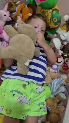 A young boy is lying on his back amidst a large collection of stuffed animals. He is wearing a blue and white striped t-shirt and green shorts with shark and star patterns. His left arm is extended, and his right arm is bent with his hand near his face. He is looking up and appears to be laughing or smiling. The stuffed animals surrounding him are of various colors and types, including a large green frog, a pink unicorn, monkeys, and bears. One stuffed animal with a brown furry texture briefly comes into the foreground, obscuring the boy's face. The setting appears to be indoors, possibly a crib or a play area filled with toys.