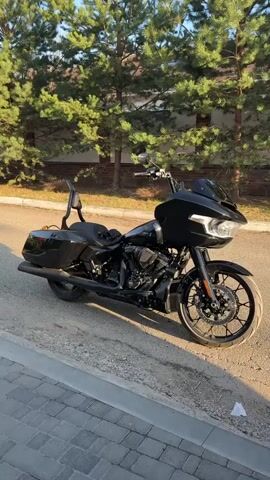 The video shows a black motorcycle parked on a paved road near a curb. The motorcycle features a front fairing, a passenger seat with a backrest, and side bags. The bike is surrounded by green trees, and the setting sun casts a warm glow on the pavement.
