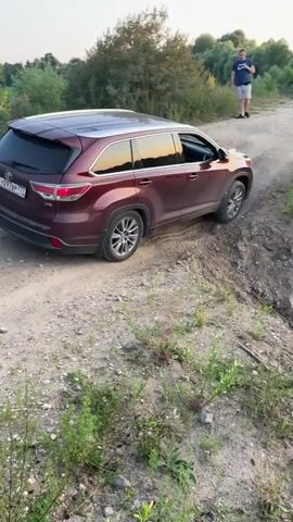 A dark red SUV is driving on a dirt road. A man is standing on the side of the road, watching. Another white SUV is parked in the background. The sun is setting, casting a warm glow over the scene. The terrain is uneven and appears to be a quarry or an undeveloped area. The dark red SUV is making a turn, kicking up dust.