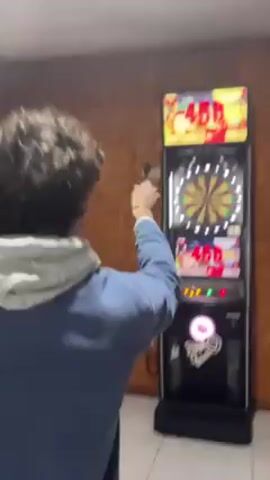 A person with curly hair, wearing a blue jacket and a white hooded sweatshirt underneath, is playing darts. They stand facing an electronic dartboard with a screen displaying the score. The dartboard machine is black and has Christmas-themed graphics on the screen. The person throws two darts during the video. The setting appears to be indoors, with a wood-paneled wall behind the dartboard.