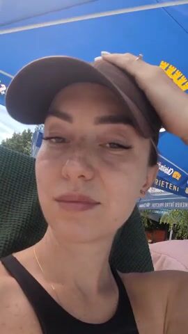A woman wearing a brown baseball cap and a black tank top is shown in a close-up video. She has freckles on her face and is wearing eyeliner and lip gloss. She is holding her hand up to the brim of her cap. She is sitting outdoors, possibly at a beach or poolside, with a blue umbrella visible in the background.