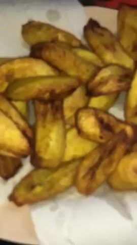 A pile of fried plantains rests on a white paper towel. The plantains are irregularly shaped, golden brown, and appear slightly oily. In the blurry background, a few red tomatoes are visible. The scene is brightly lit.